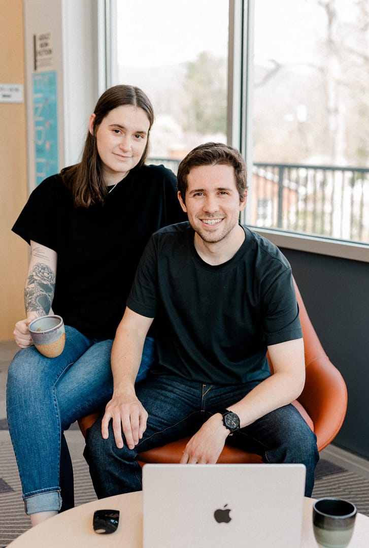 Owners of Barefoot Build sit next to each other on an orange chair, next to a computer and mouse.